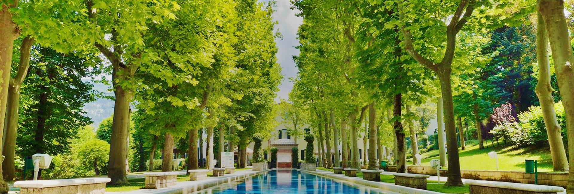 Viale alberato con piscina riflettente, circondato da panchine e verde lussureggiante.